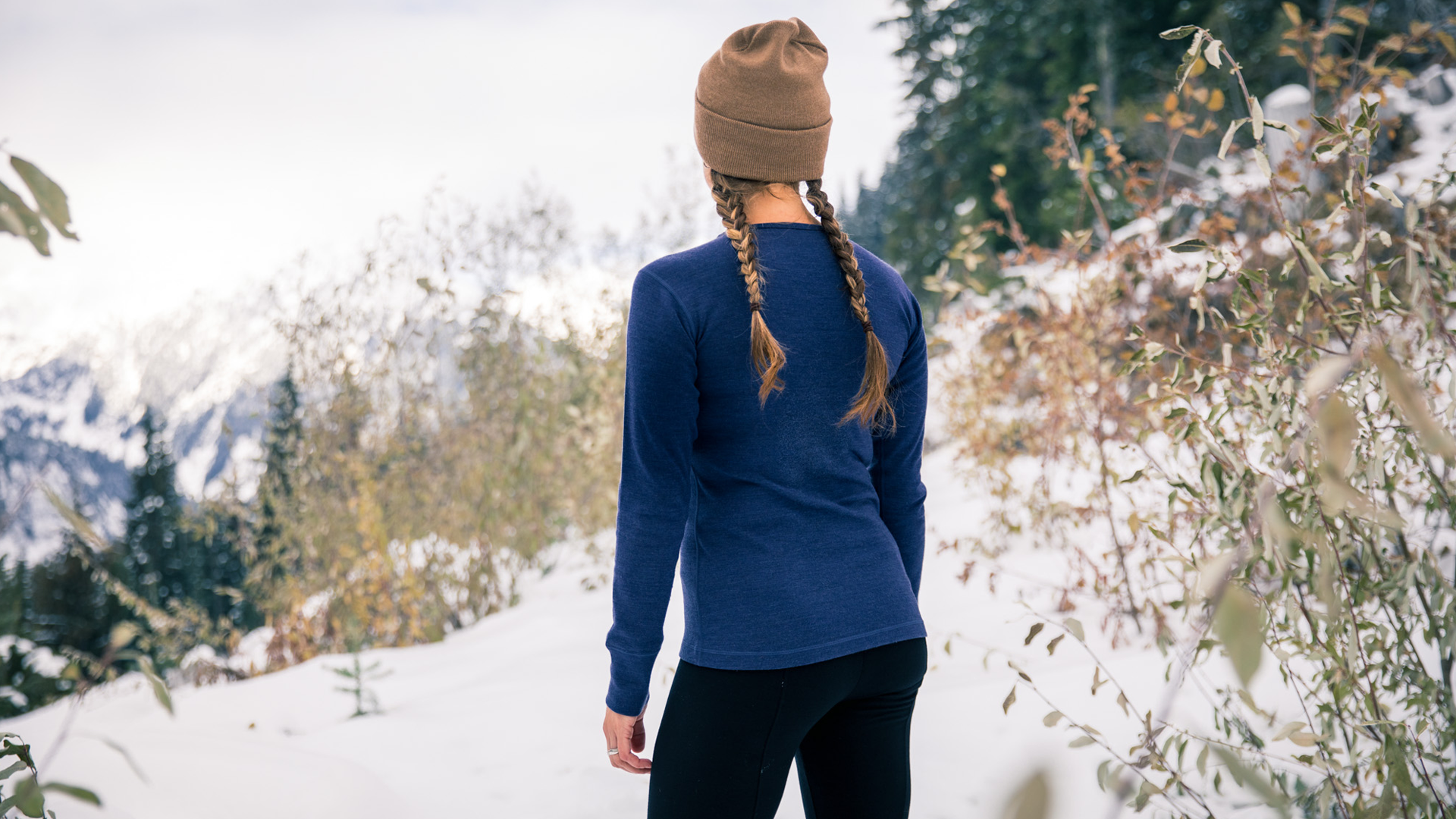 A person with braided hair, wearing a brown beanie, a blue long-sleeve shirt, and black leggings, stands in a snowy landscape with mountains and trees in the background, looking into the distance.