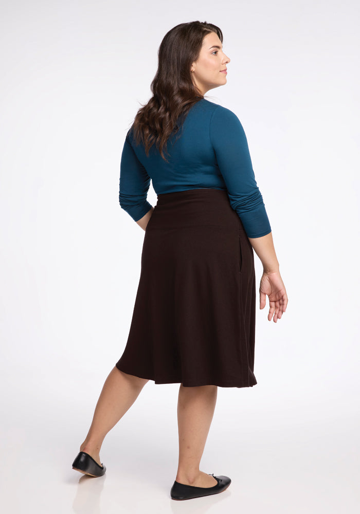 A woman with long, dark hair stands facing away, wearing a blue long-sleeve top and the Marilyn Flowy Midi Skirt in French Roast by Woolx, featuring an A-line silhouette. She completes the ensemble with black flats against a plain white background.