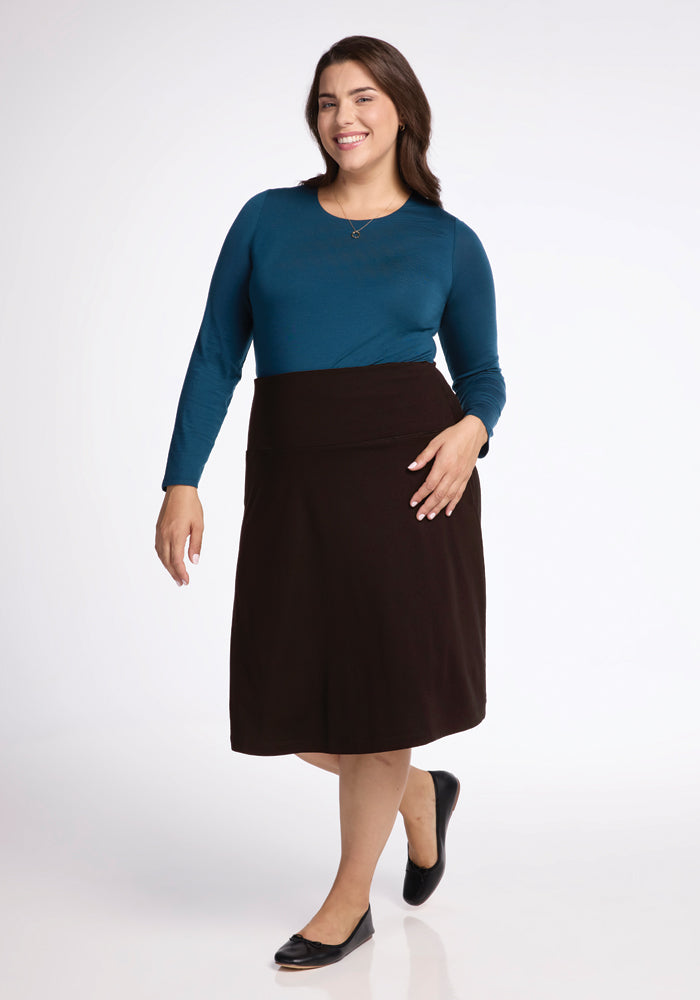 Dressed in a teal long-sleeve shirt, a person poses with a smile while showcasing the Woolx Marilyn Flowy Midi Skirt in French Roast. Their long hair adds to the sophisticated A-line silhouette of the outfit, which they finish off with black flats against a plain white backdrop.