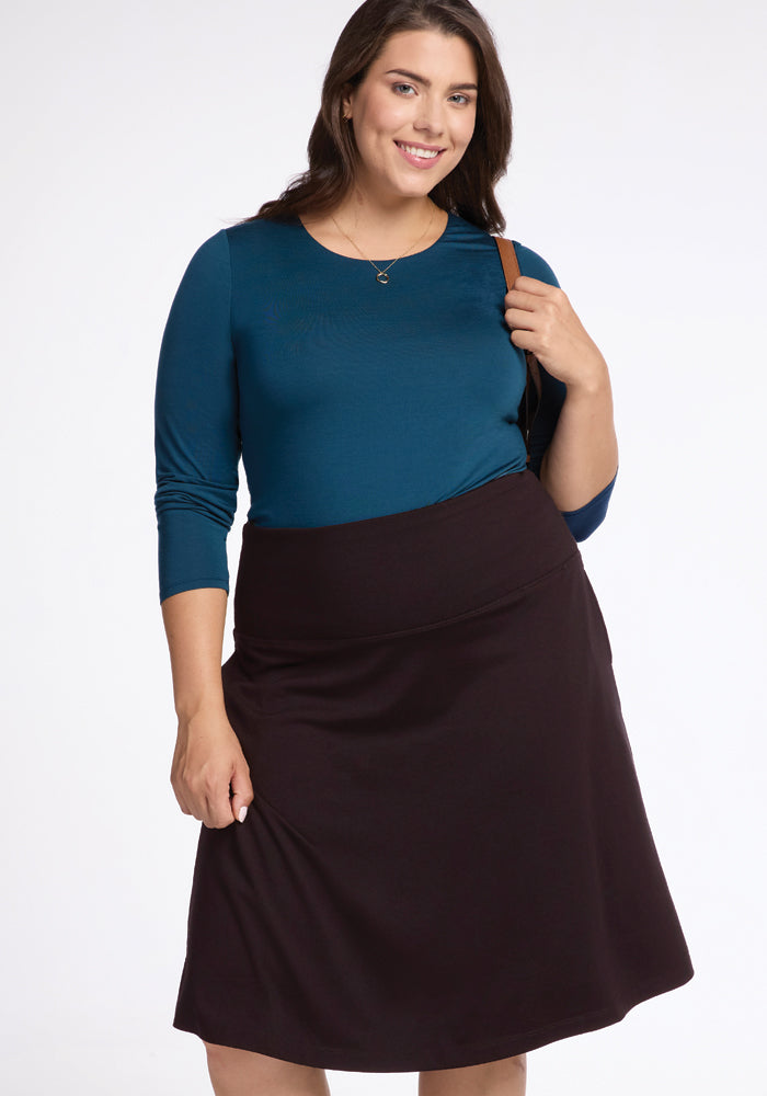 A smiling woman stands against a plain white background, wearing a teal long-sleeve top paired with the Marilyn Flowy Midi Skirt in French Roast by Woolx. She holds the strap of a bag on her shoulder, exuding effortless style.