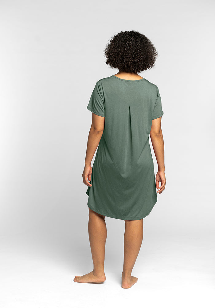 A person wearing the Woolx Desi Nightgown OLD, featuring a short-sleeve design in a knee-length green fabric reminiscent of comfortable sleepwear, stands barefoot on a white background with their back turned. The gown highlights pleated detailing at the back.