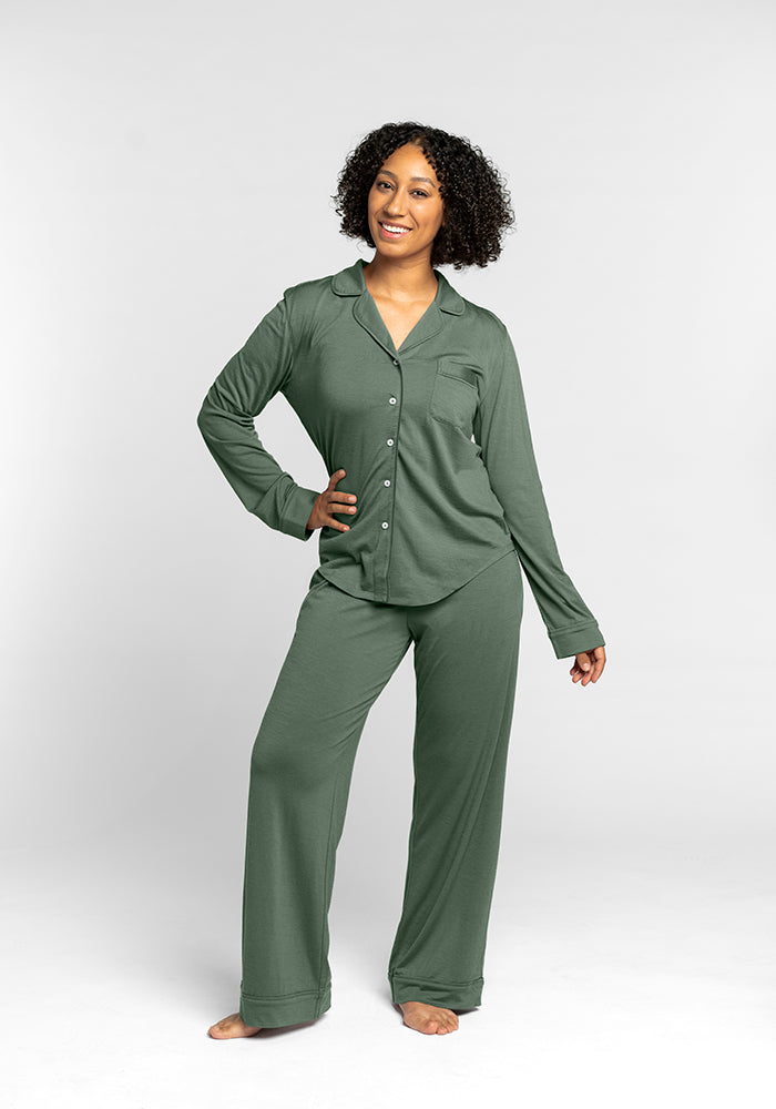 A person smiles, wearing the Harper FeatherTouch® Pajama Set by Woolx in Duck Green, made from temperature-regulating Merino wool. The set includes a button-up long-sleeve top and pants. They stand barefoot against a gray background, highlighting the comfort of this cooling sleepwear.