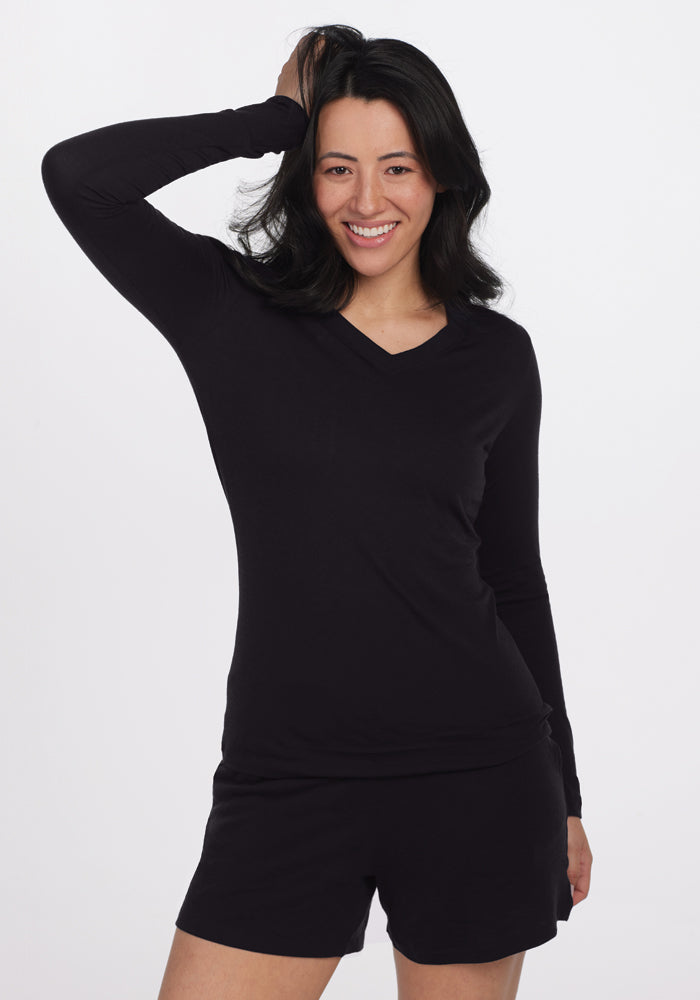 A person with long dark hair smiles at the camera, wearing a cozy Woolx Lily V Neck Lounge Top in black and black shorts. They pose confidently with one hand resting on their head against a plain white background.