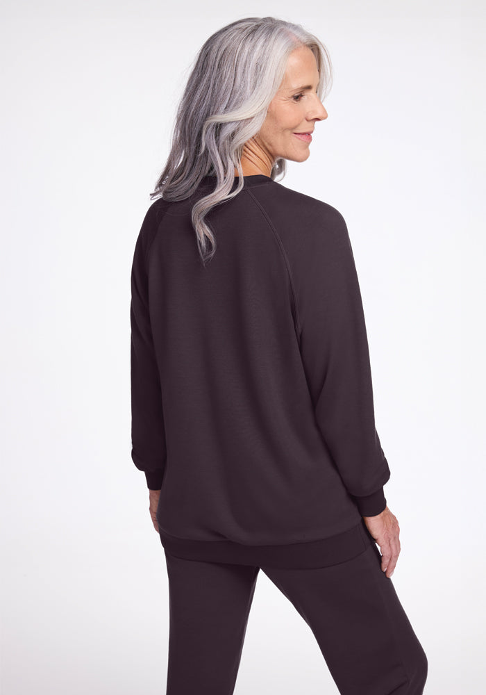 An older adult with long, gray hair stands facing away, dressed in a dark casual outfit from Woolx. The Bailey Crewneck Sweatshirt in Chocolate Plum, made with sustainable Merino wool, is set against a plain and light background, accentuating the relaxed and comfortable style of the attire.