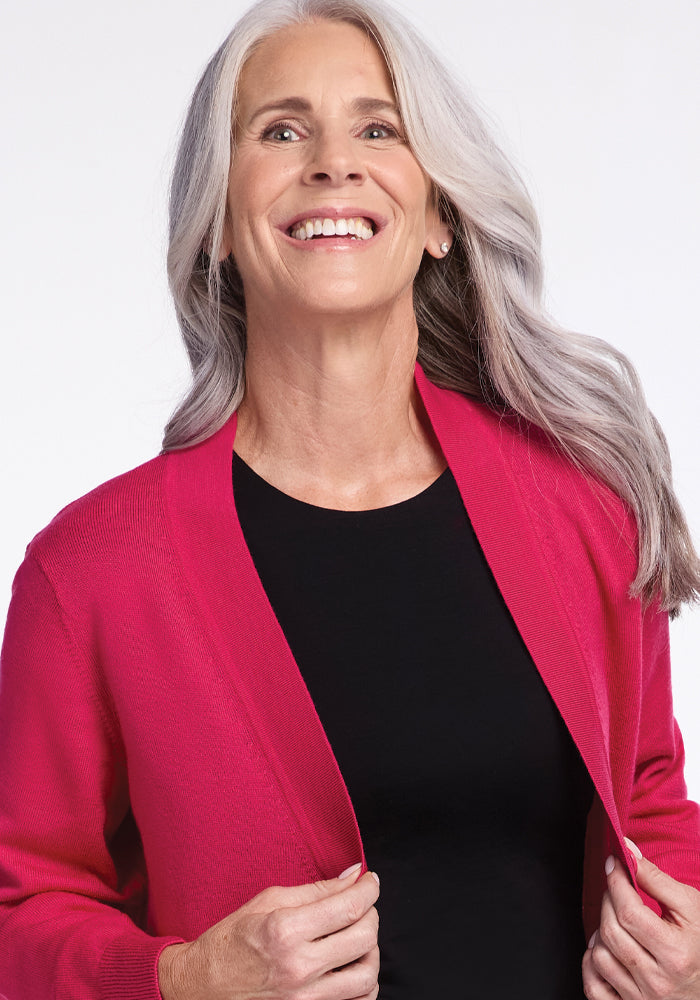 A smiling woman with long gray hair wears the Gia Sweater in Berry Burst from Woolx over a black shirt. She is looking upward, holding the edges of her sweater against a plain white background.