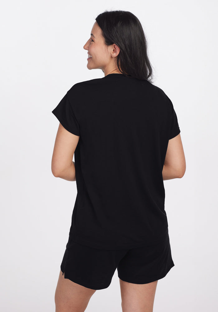 A person with long, dark hair is standing and smiling while looking over their shoulder. They're wearing the Billie Lounge Tee in black by Woolx, known for its temperature-regulating and moisture-wicking properties, set against a plain white background, creating an impression of effortless comfort and style.