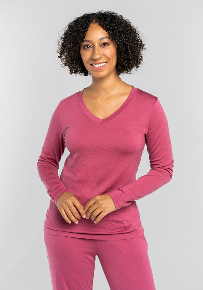 A person with curly hair smiles while wearing Woolx's Lily V Neck Lounge Top in Red Violet, paired with matching pants made of moisture-wicking Merino wool. The background is a plain light gray.