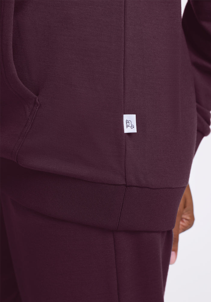 Close-up of a person wearing the Avery Hoodie in Wine Tasting from Woolx, showcasing part of the hoodie and matching pants. A small white tag with a minimal graphic is visible on the side of the hoodie against a plain white background.