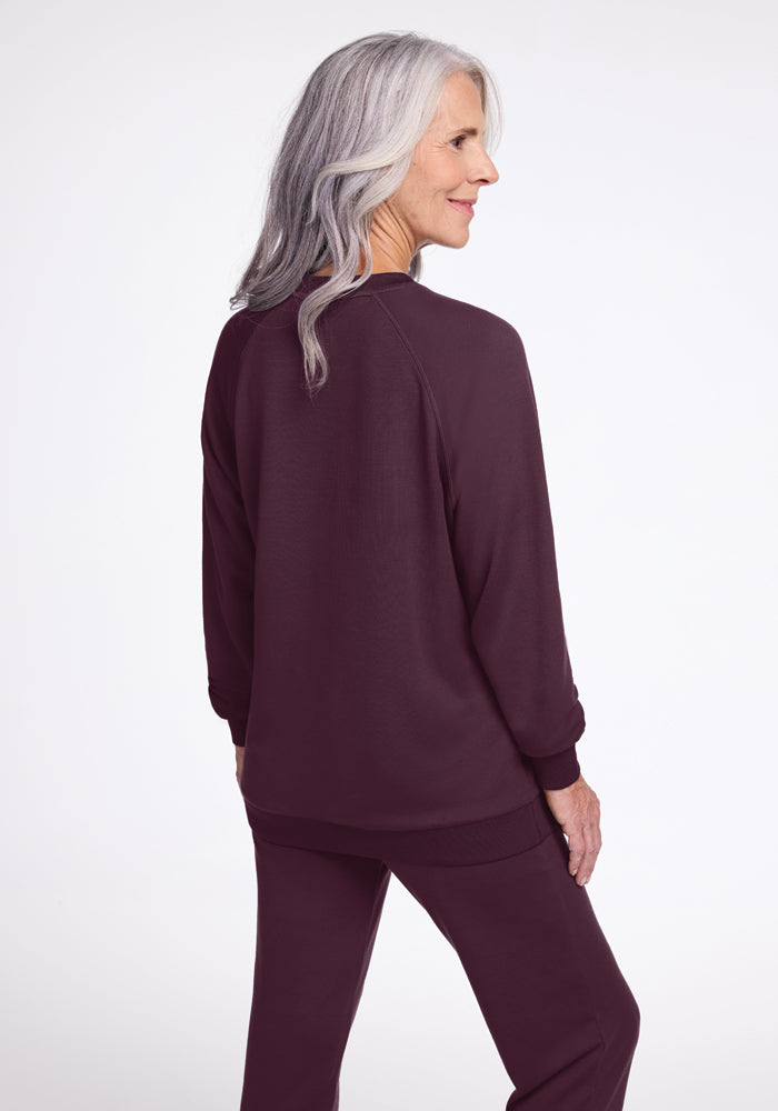 A woman with long gray hair is smiling while standing and looking to the side. She is wearing a matching maroon sweatshirt and pants, featuring the Bailey Crewneck Sweatshirt in Wine Tasting from Woolx's sustainable collection. The background is plain white.