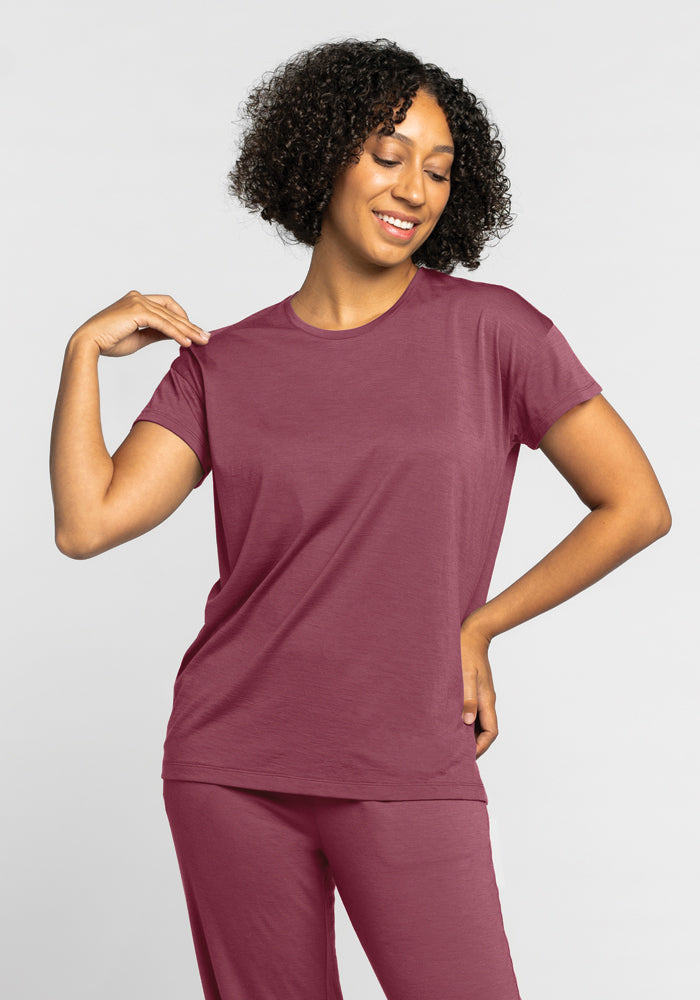 A person with curly hair is smiling, dressed in the temperature-regulating Billie Lounge Tee in Wild Ginger from Woolx. They pose with one hand on their shirt collar and the other on their hip, set against a plain light gray background. 