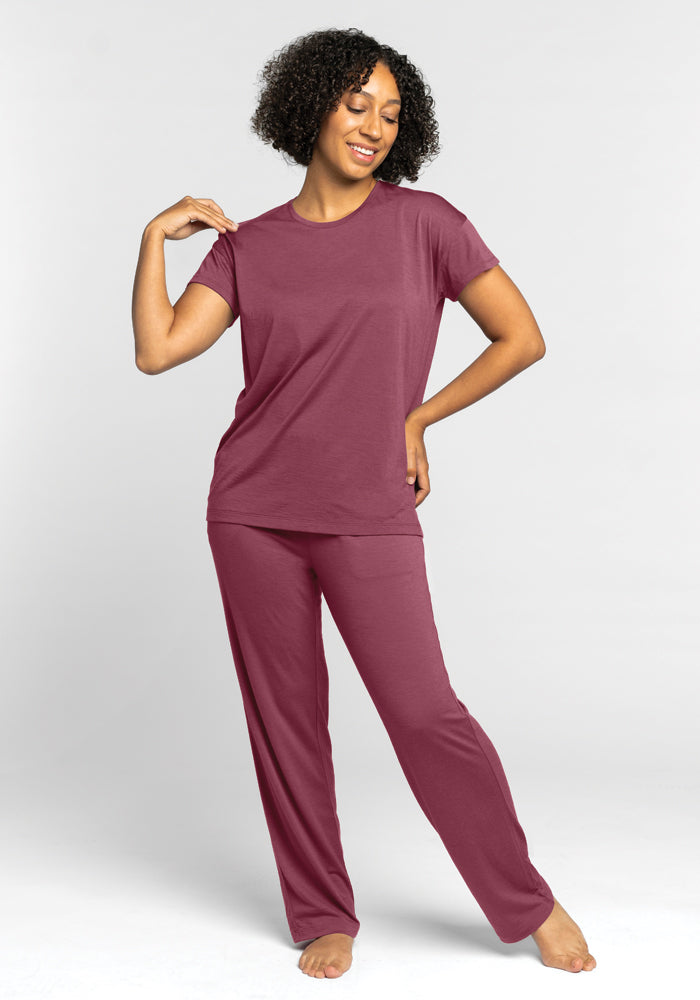 A person stands barefoot, smiling with closed eyes while wearing the Woolx Billie Lounge Tee in Wild Ginger. They pose with one hand on their hip and the other slightly raised. The tee's temperature-regulating fabric keeps them perfectly at ease against a plain, light-colored background.