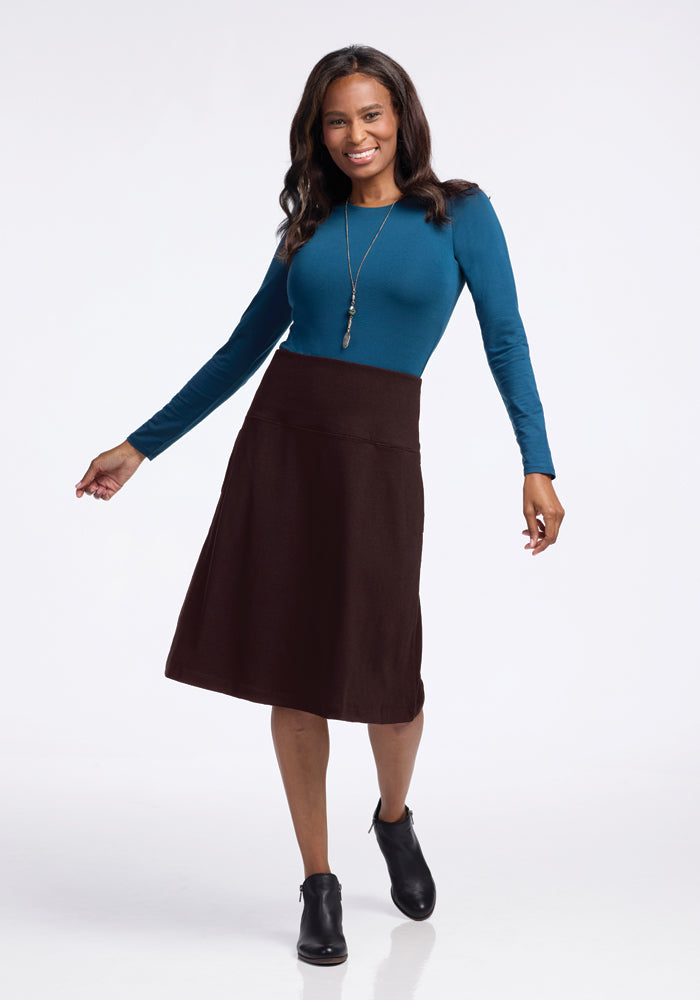 A person smiling, wearing a teal long-sleeve top, the Marilyn Flowy Midi Skirt in French Roast by Woolx with an A-line silhouette, and black ankle boots. They have long hair and are standing on a light background. A pendant necklace completes the look.