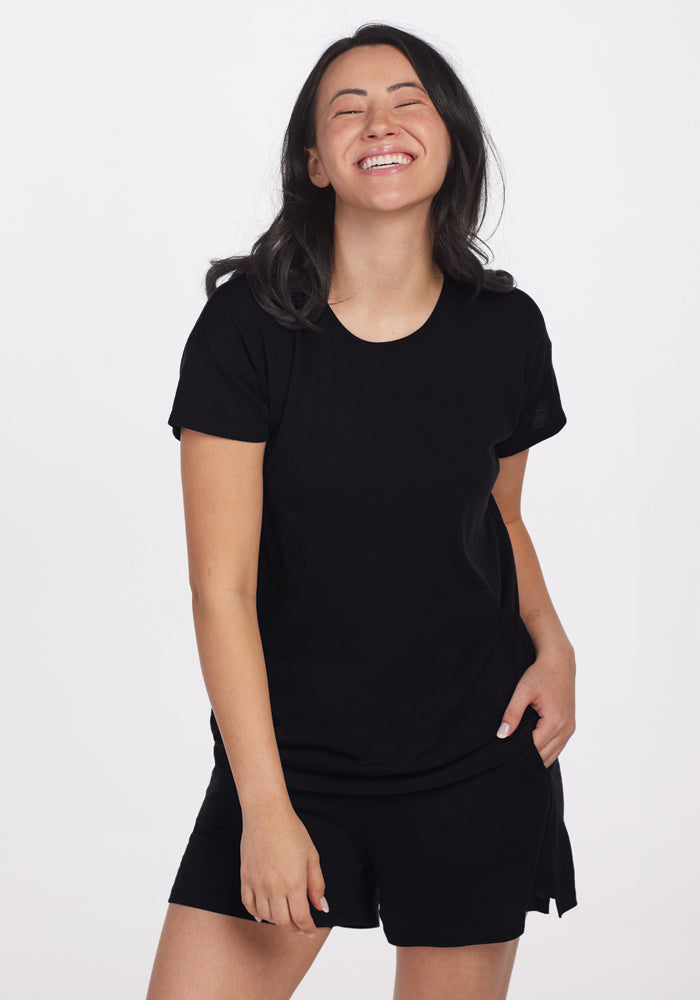 A person with long black hair is smiling and wearing the stylish Billie Lounge Tee in black and shorts, standing against a plain white background with one hand in their pocket. The outfit suggests temperature-regulating comfort that brings to mind cozy merino wool pajamas, courtesy of Woolx.