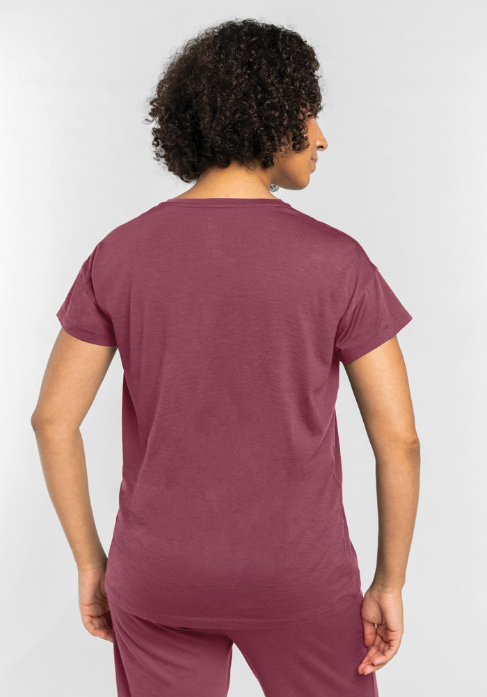 A person with curly hair stands facing away from the camera, wearing a Woolx Billie Lounge Tee in Wild Ginger and matching moisture-wicking pants, set against a plain backdrop.