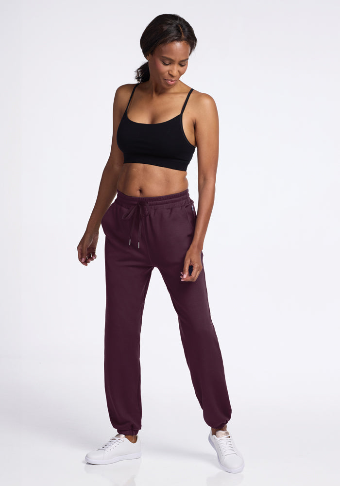 A person wearing a black sports bra, Parker Sweatpants in Wine Tasting from Woolx, and white sneakers stands against a plain white background. The individual gazes downwards, with one hand in a pocket, showcasing a relaxed and athletic stance.