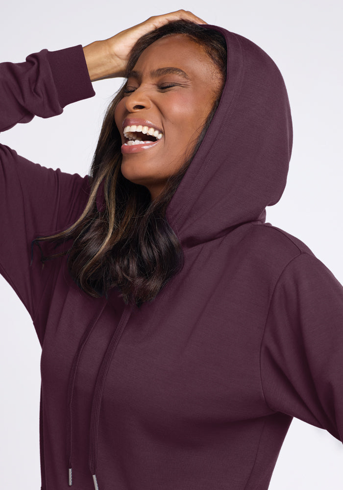 A woman wearing the Woolx Avery Hoodie in Wine Tasting, with a cozy kangaroo pouch, is smiling and looking upwards with one hand resting on her head. She has long, wavy hair and stands against a light background.