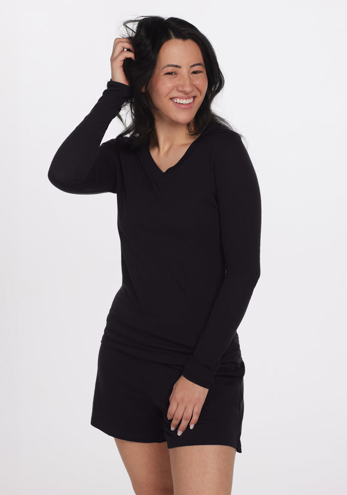 A person with long dark hair smiles while wearing a black long-sleeve shirt similar to the Woolx Lily V Neck Lounge Top, paired with black shorts. They stand against a plain white background, one hand gently touching their hair.