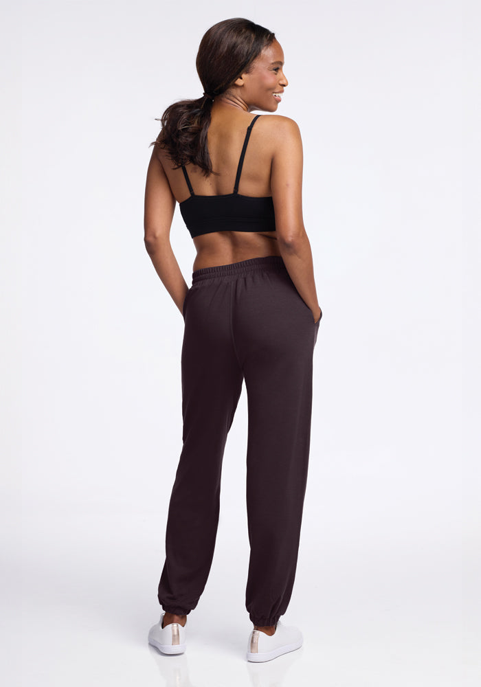 A woman stands with her back to the camera, wearing a black sports bra and Woolx's Parker Sweatpants in Chocolate Plum. She has her hands in her pockets and is smiling. Her hair is in a ponytail, and she sports white sneakers. In these odor-free, temperature-regulating pants, she strides confidently against a plain white backdrop.