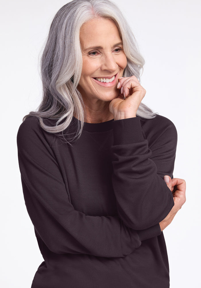 An older woman with long, wavy gray hair and a warm smile is posed with her hand touching her cheek. She is wearing the Woolx Bailey Crewneck Sweatshirt in Chocolate Plum against a plain light background.