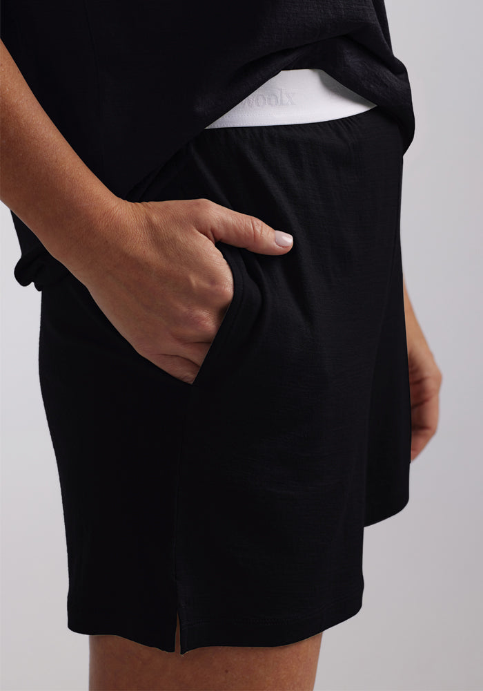 A person wearing the Poppy Lounge Shorts in black, crafted from cozy Merino wool by Woolx, stands against a light gray background. With one hand in the shorts pocket, the waistband of white undergarments is slightly visible above these comfortable shorts, hinting at a peaceful night's sleep.