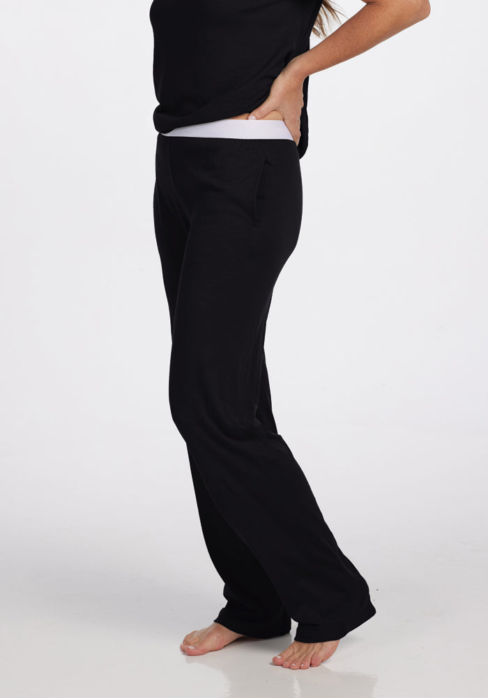 A person wearing black Maya Lounge Pants by Woolx and a matching top stands barefoot on a white background. With one hand on their hip and the other arm extended downward, only the lower half of their face is visible, while the comfortable waistband complements the sleek look.