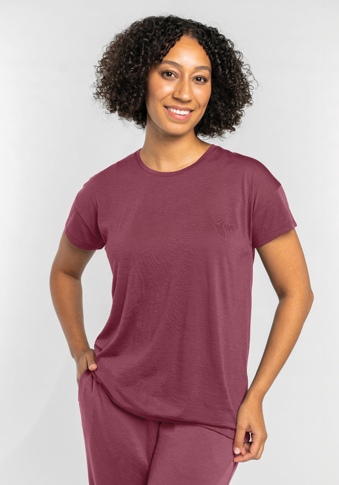 A person with curly hair smiles while wearing a matching Billie Lounge Tee in Wild Ginger by Woolx, along with pants made of moisture-wicking fabric. They stand against a plain light background, with one hand resting on their hip.