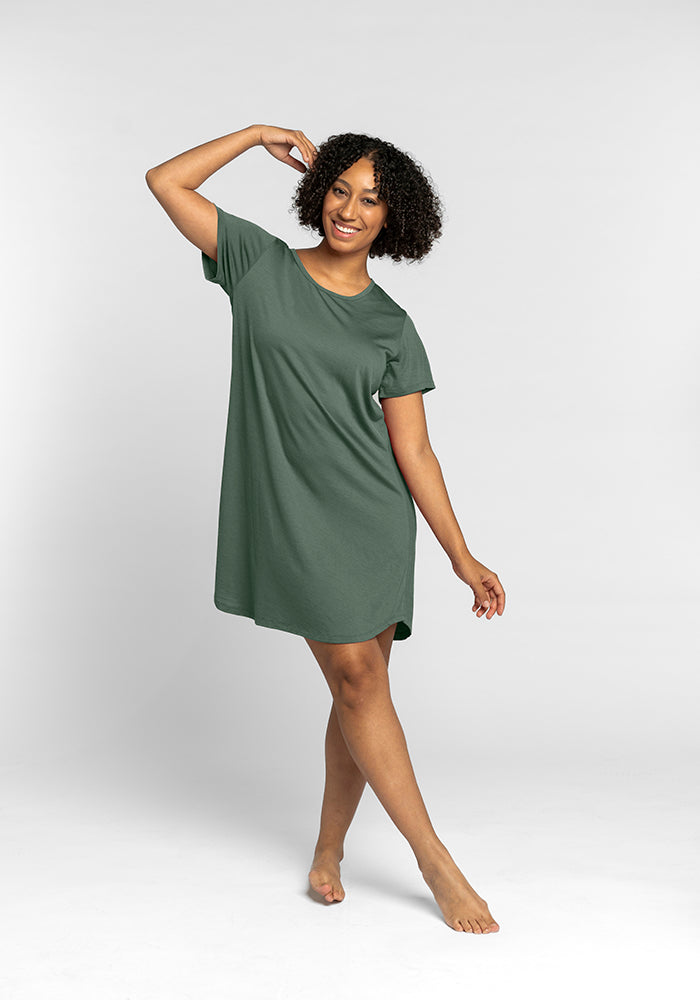 A person with curly hair smiles while posing in a loose, short-sleeved green dress from Woolx, known as the Desi Nightgown OLD. They balance gracefully on one foot against a plain white background, exuding comfort and style.