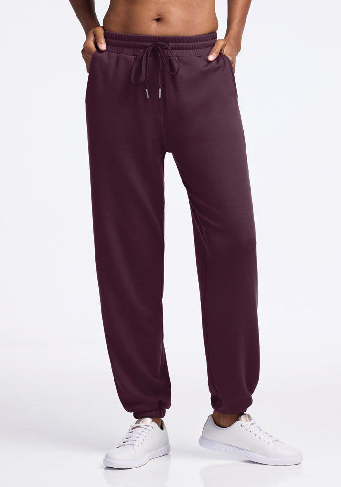 A person is wearing Woolx's Parker Sweatpants in Wine Tasting color with white sneakers, standing against a plain white background. These sweatpants provide travel comfort with features like a drawstring waist, elastic cuffs, and moisture-wicking properties to keep you cozy on the go. 