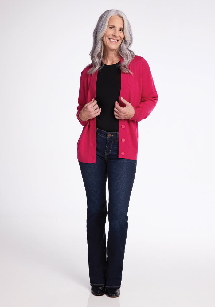 A woman with long gray hair stands smiling against a plain white background, wearing a black top, dark jeans, and the Woolx Gia Sweater in Berry Burst.