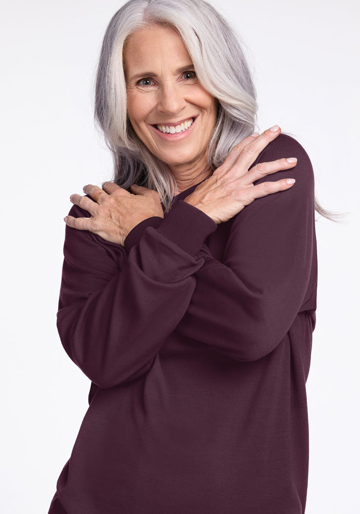 A smiling older woman with long gray hair embraces herself while wearing the Bailey Crewneck Sweatshirt in Wine Tasting from Woolx. This sustainable sweatshirt stands out against the plain white background, offering warmth and comfort with its Merino wool blend.