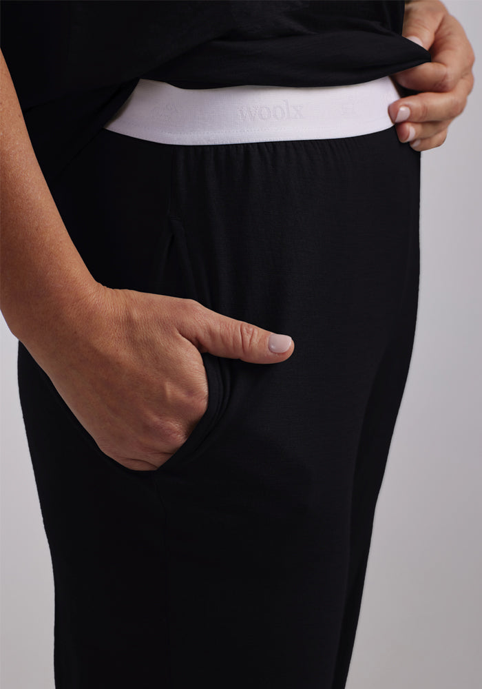 A person is depicted from the side, wearing black Maya Lounge Pants by Woolx, which feature a snug white waistband. The brand's name "Woolx" is visible on the waistband, emphasizing its signature Merino wool blend. Their hand rests in the pocket for a relaxed look.