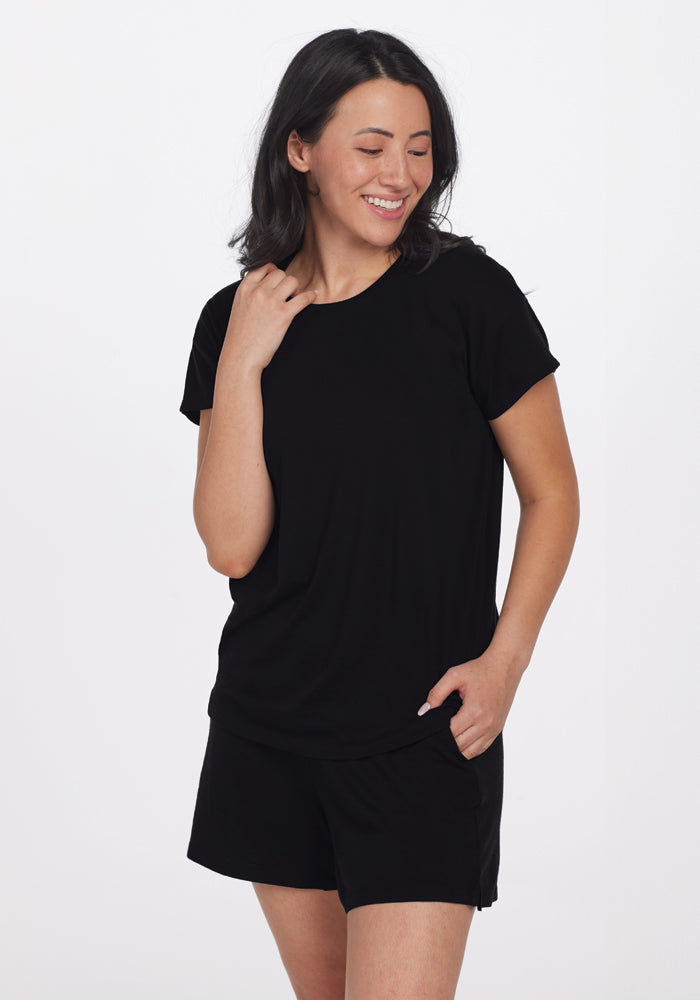 A smiling person with shoulder-length dark hair is wearing the Billie Lounge Tee in black from Woolx, along with black shorts made from moisture-wicking fabric. They stand against a plain white background, one hand on their neck and the other in their pocket.