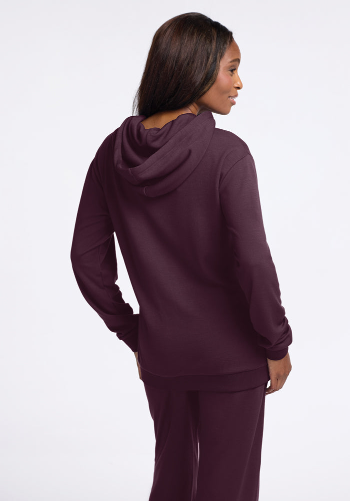 A woman in the Woolx Avery Hoodie - Wine Tasting, complete with a kangaroo pouch and matching pants, stands against a white background. She faces away from the camera, her profile visible, showcasing the back of the hoodie and her long, straight hair.