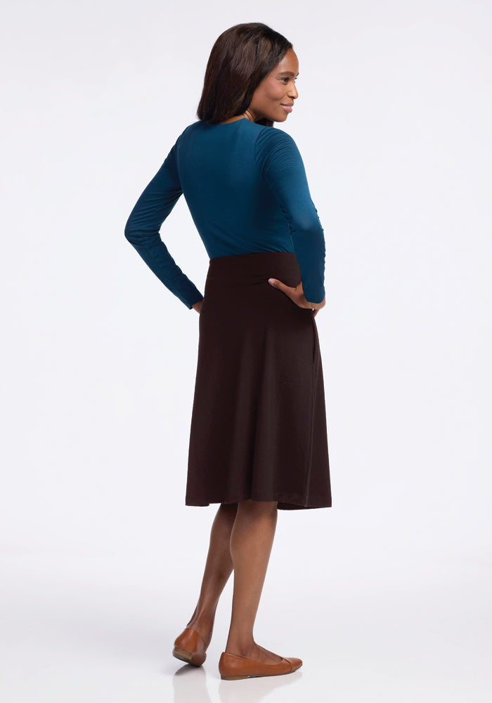 A woman stands with her back to the camera, wearing a teal long-sleeve top paired with the Woolx Marilyn Flowy Midi Skirt in French Roast, which elegantly embraces an A-line silhouette. Her brown flats complement the ensemble as she confidently places her hands on her hips against a plain white background.