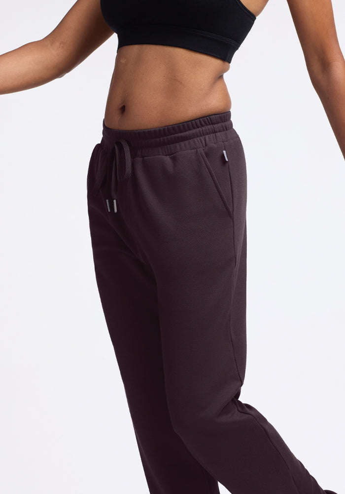 A person wearing a black crop top and Woolx Parker Sweatpants in Chocolate Plum poses against a plain white background. With arms slightly extended, the focus is on the midsection and clothing, emphasizing the stylish outfit's temperature-regulating features.