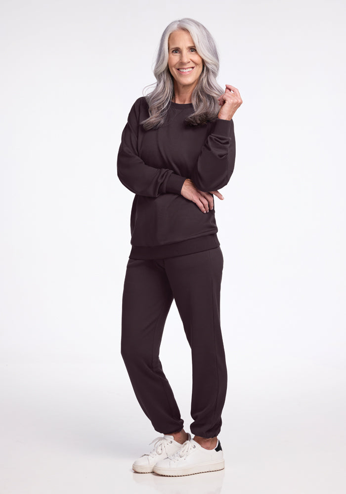 A smiling woman with long gray hair stands confidently against a plain background. She's wearing a sleek chocolate plum Bailey Crewneck Sweatshirt from Woolx, paired with matching pants and white sneakers. Her right hand lightly touches her hair as her left arm rests across her torso, exuding effortless style.