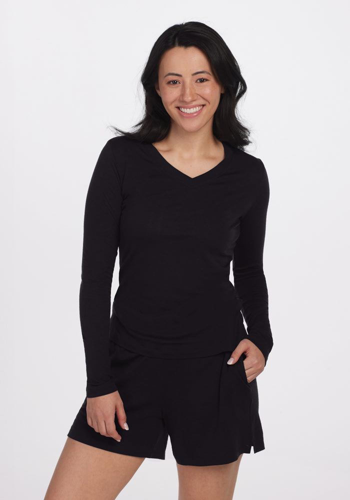 A person with long dark hair is smiling, wearing a Woolx Lily V Neck Lounge Top in black and black shorts. They stand comfortably against a plain white background. 