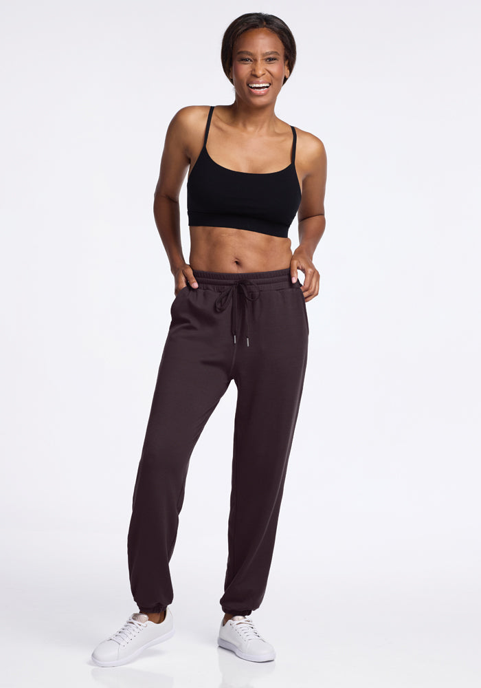 A person wearing a black sports bra, Parker Sweatpants in Chocolate Plum by Woolx, and white sneakers stands against a plain background, smiling with hands on hips. The temperature-regulating and odor-free Merino wool fabric ensures comfort and freshness.