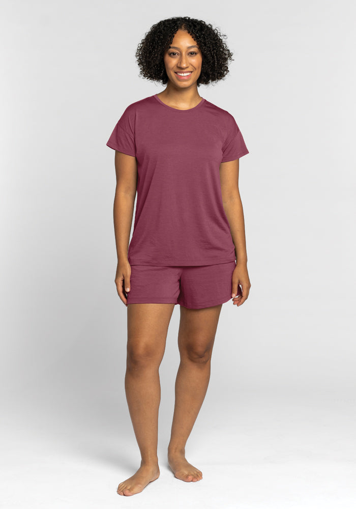 A person wearing the Billie Lounge Tee in Wild Ginger by Woolx, along with matching shorts crafted from moisture-wicking fabric, stands barefoot against a plain, light gray background. With curly hair and a warm smile, they exude comfort and style effortlessly for the camera.