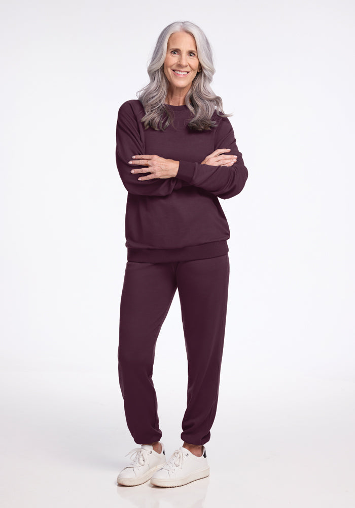 A person with long gray hair stands confidently, arms crossed, wearing a maroon tracksuit and white sneakers. The outfit features the Bailey Crewneck Sweatshirt in Wine Tasting from Woolx's sustainable Wool Aire Collection, made of Merino wool and set against a plain white background.