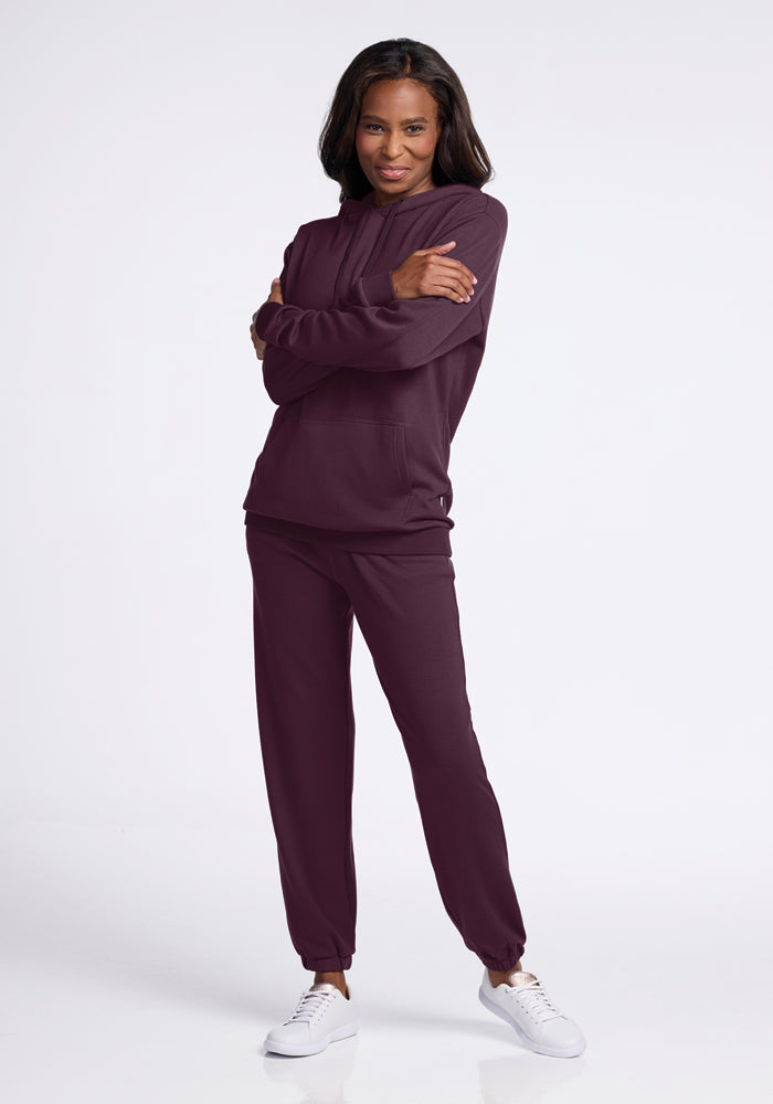 A person stands confidently with arms crossed, wearing the Avery Hoodie in Wine Tasting by Woolx, paired with white sneakers. The hoodie features a kangaroo pouch pocket, perfectly blending athletic style and casual comfort against the plain white background.