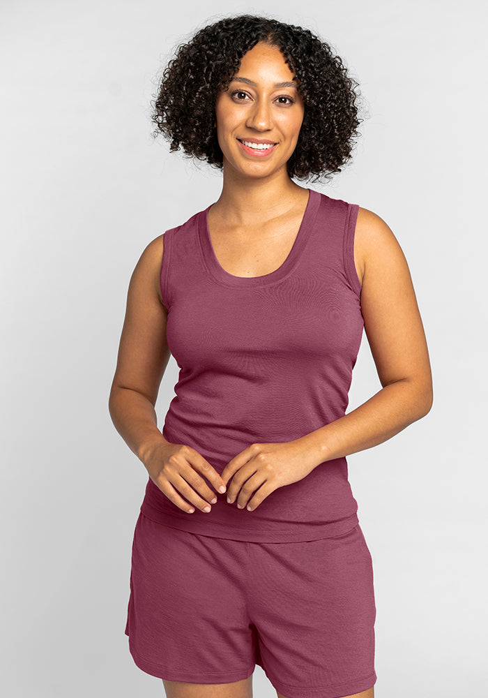 A person with curly hair smiles while wearing a matching burgundy sleeveless top and shorts set, reminiscent of the Woolx Phoebe Lounge Tank in Wild Ginger. The outfit is both odor-free and temperature-regulating, perfect for comfort. The background is a plain light gray. 