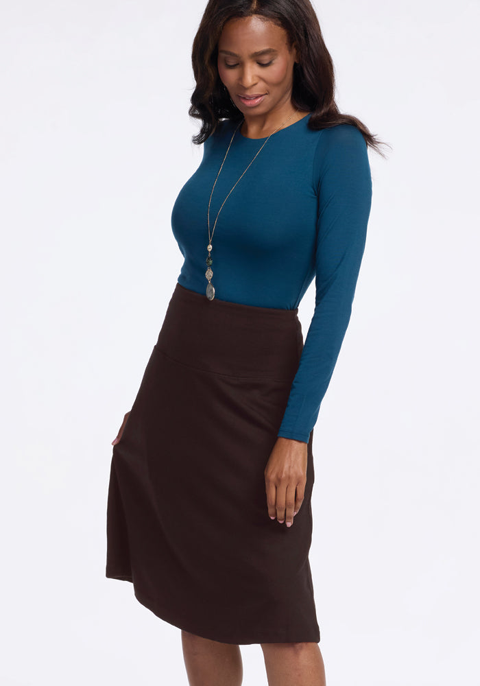 A woman with long hair stands smiling, wearing a teal long-sleeve top paired with the Marilyn Flowy Midi Skirt in French Roast by Woolx, along with a long necklace. The background is plain white.