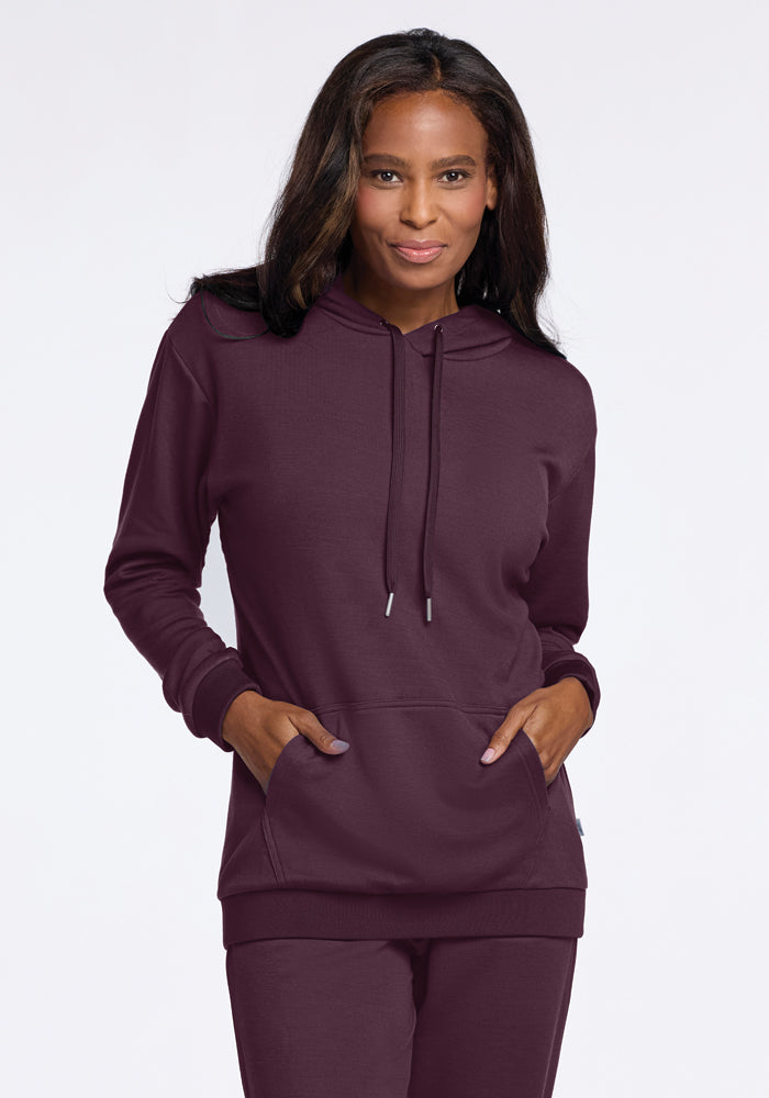 A person dressed in a Wine Tasting Avery Hoodie from Woolx stands against a plain background, smiling at the camera. Made from soft Merino wool, they have their hands casually tucked into the front pocket of the hoodie.