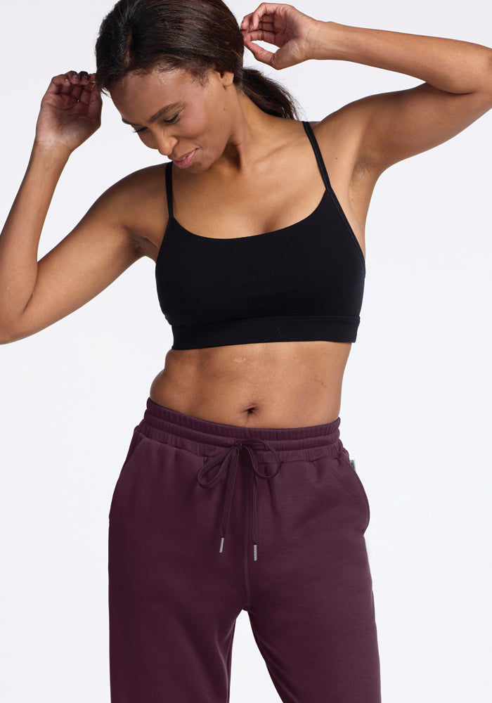 A person dressed in a black sports bra and Woolx's Parker Sweatpants in Wine Tasting, which are recognized for their odor-free and temperature-regulating Merino wool, is posing with arms up, looking down, and smiling against a plain white background.