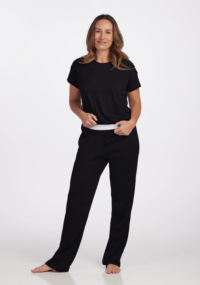 A person stands barefoot on a plain background, wearing a casual black ensemble that includes Woolx's Maya Lounge Pants, which feature a comfortable waistband. They pair it with a short-sleeved top, resting their hands in their pockets with a relaxed expression.