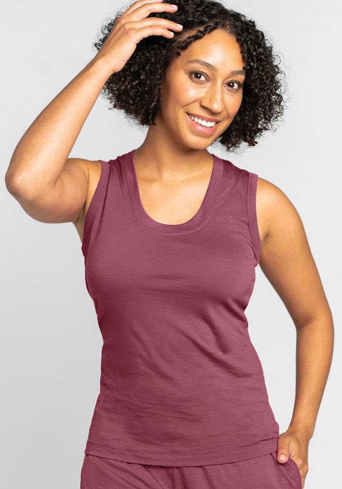 A person with curly hair smiles while holding one hand up to their head. They are wearing the temperature-regulating, sleeveless Phoebe Lounge Tank by Woolx and matching bottoms in Wild Ginger. The background is plain and light-colored.