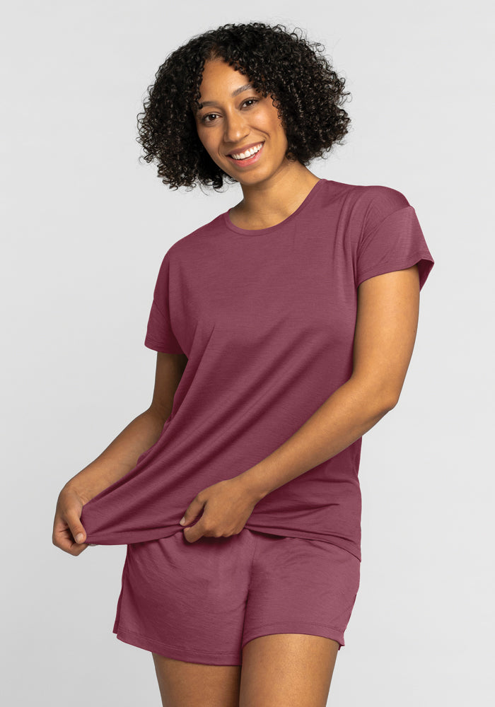 A person with curly hair smiles, wearing the Billie Lounge Tee in Wild Ginger by Woolx, along with matching shorts. Made from moisture-wicking fabric, they gently hold the hem of the shirt with one hand against a plain, light-colored background that highlights their relaxed style.