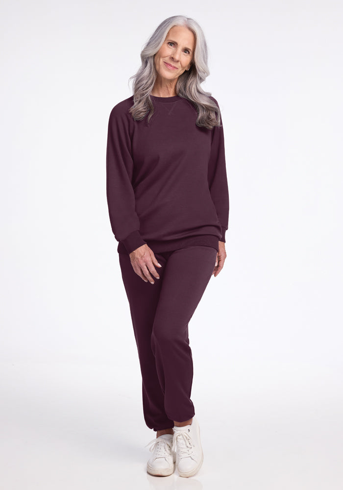 An older woman with long gray hair stands in a relaxed pose, showcasing the Bailey Crewneck Sweatshirt in Wine Tasting from Woolx's sustainable collection. She pairs it with matching maroon sweatpants and white sneakers against a plain white background.