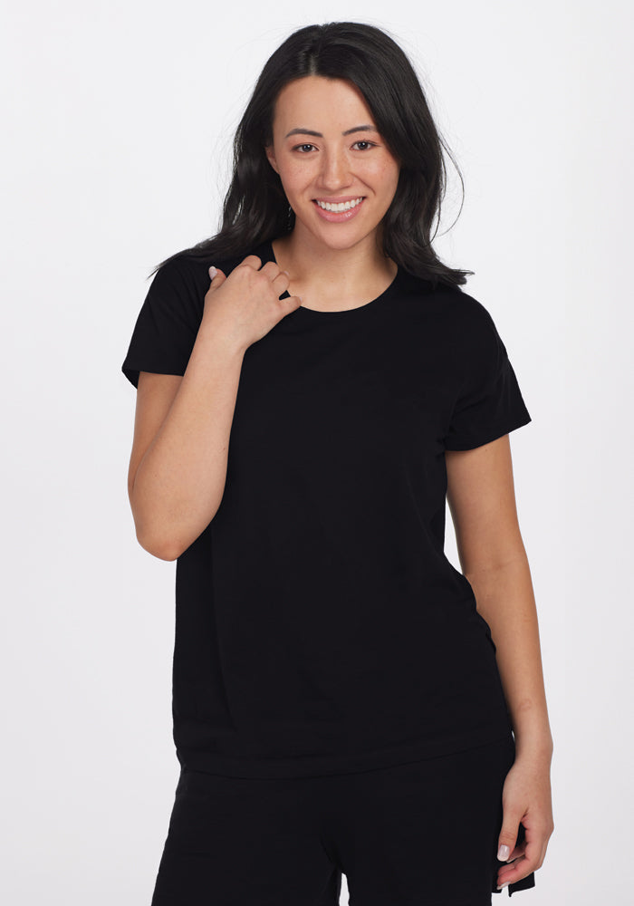 A woman with long, dark hair, wearing the Billie Lounge Tee in black from Woolx designed for moisture-wicking comfort, smiles and poses against a plain white background. 